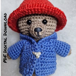 May include: Crocheted teddy bear wearing a red hat and a blue coat with a toggle closure. The bear is brown with black eyes and a black nose. The text "PDF DIGITAL DOWNLOAD" is on the left side of the image.