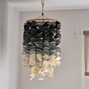 Può includere: Una lampada a sospensione con una cornice circolare intrecciata e una cascata di dischi circolari a strati. I dischi cambiano colore dal grigio scuro nella parte superiore al crema chiaro nella parte inferiore, creando un effetto sfumato. La lampada è appesa a un cordoncino in fibra naturale.