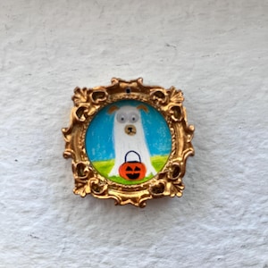 May include: A small, square, ornate gold-colored frame containing a circular painting of a white dog wearing a ghost costume. The dog is positioned on a green field against a blue sky and is holding an orange jack-o'-lantern candy bucket.