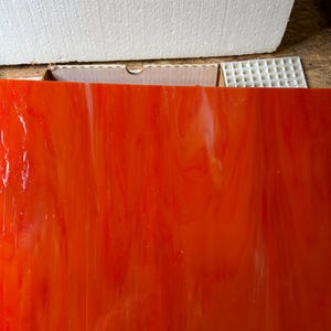 May include: A sheet of vibrant orange stained glass with a flowing, marbled appearance. The glass has a glossy finish and is illuminated, showcasing the color variations. The sheet is rectangular and appears to be part of a larger project.