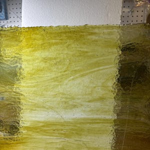 May include: A sheet of textured, translucent stained glass in shades of yellow and gold. The glass has a wavy, uneven edge and a marbled appearance, with streaks and swirls of color throughout. The light passes through the glass, creating a warm, inviting glow.