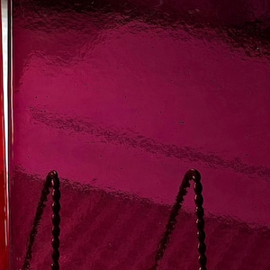 May include: A close-up of a shiny, deep magenta glass object with a textured surface. Two dark, twisted metal supports are visible at the bottom. The object has a reflective quality, with highlights and shadows.