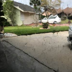 May include: A view of a suburban neighborhood through a window covered in raindrops. The houses are visible through the water droplets, and a white car is parked in the driveway.