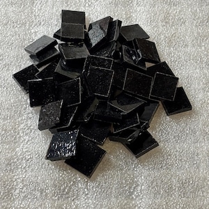 May include: A pile of small, square, black glass tiles. The tiles have a glossy, reflective surface and are scattered on a textured, light-colored surface. The tiles appear to be for mosaic art or craft projects.