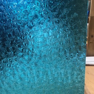 May include: A close-up of a teal blue glass panel with a textured pattern of small, raised circles. The glass has a gradient effect, with the top being a darker shade of blue and the bottom being a lighter shade.