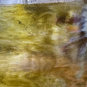 May include: A sheet of textured stained glass with a blend of golden yellow and brown hues. The glass has a rippled, flowing appearance, with light reflecting off the surface. The glass is likely for crafting or decorative purposes.