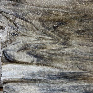 May include: A sheet of textured art glass with a swirling pattern of white, grey, and brown. The glass has a rippled surface and a speckled, glittery appearance, creating a unique visual texture. The overall effect is reminiscent of a stormy sky or marbled stone.