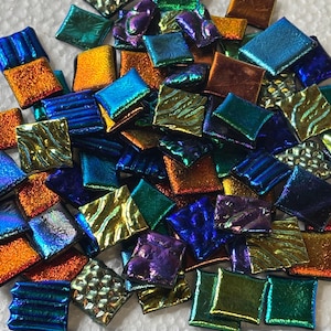 May include: A collection of small, square glass tiles in various iridescent colors, including blue, green, orange, and purple. The tiles are arranged in a pile, with a ruler in the foreground for scale. Each tile is approximately 0.5 inches square.