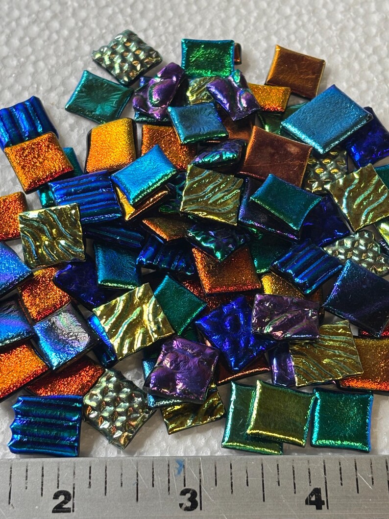 May include: A collection of small, square glass tiles in various iridescent colors, including blue, green, orange, and purple. The tiles are arranged in a pile, with a ruler in the foreground for scale. These tiles are suitable for mosaic art projects.