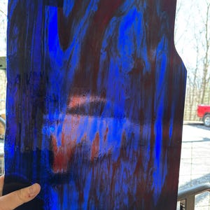 May include: A rectangular sheet of stained glass with a vibrant blend of blue, black, and red hues. The glass has a unique, flowing pattern, with a dark blue border and a lighter blue center. The edges are uneven, and there is a small, rectangular cutout at the top.