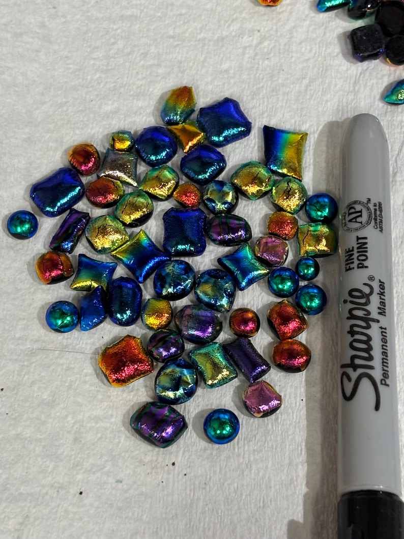 May include: A collection of iridescent glass pieces in various shapes and sizes, including squares, circles, and irregular forms. The colors shift from blue to gold, red, and purple. A gray Sharpie marker is included for scale.