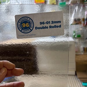 May include: A sheet of clear, double-rolled glass with a textured surface. A sticker on the glass reads "Paul Wissmach Glass Co. 96-01 2mm Double Rolled". The glass is held by a hand.