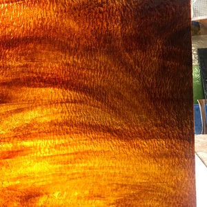 May include: A textured, amber-colored glass panel with a rippled surface. The glass has a warm, golden hue with darker brown accents, creating a dynamic, light-catching effect. The panel's surface is uneven, adding depth and visual interest.