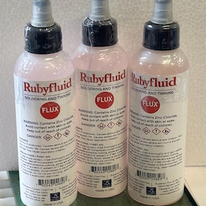 May include: Three clear plastic squeeze bottles of Rubyfluid soldering and tinning flux. Each bottle features a black cap, a red circular label with the word FLUX, and white labels containing safety warnings, hazard symbols, and barcodes.