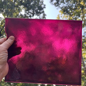 May include: A rectangular sheet of translucent magenta glass with a textured, rippled surface. The glass is held up against a bright outdoor background, allowing light to pass through and highlight the deep pink and purple color variations.