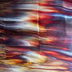 May include: A sheet of stained glass featuring a swirling, marbled pattern of deep crimson, burnt orange, golden yellow, and dark charcoal gray. The surface displays a smooth, glossy texture with translucent and opaque color variations throughout.