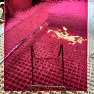 May include: A vibrant, fuchsia-colored, square glass piece with a textured surface, displayed on a decorative metal stand. The stand has a twisted wire design. The glass is resting on a white grid.