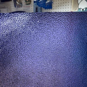 May include: Close-up of a textured, purple glass sheet. The surface is covered in small, rounded bumps, creating a unique pattern. The color transitions from a lighter shade at the top to a darker hue at the bottom, adding depth.