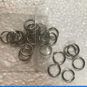 May include: A collection of small, silver-colored split rings, some open and some closed, are scattered on a white surface. A clear plastic tube containing additional rings is also present. These metal rings are likely used for jewelry making or crafts.