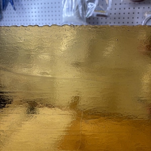 May include: Close-up of a sheet of gold leaf with a textured surface. The gold leaf has a reflective quality, with subtle variations in color and tone. The edges of the leaf appear slightly uneven.
