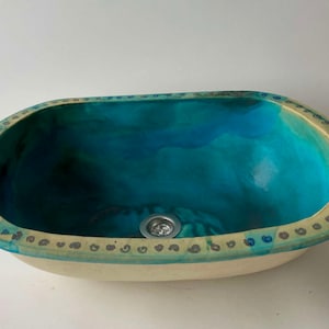 Può includere: Lavabo ovale in ceramica con smalto blu e verde e bordo bianco. Il lavabo ha un piccolo foro di scarico al centro.