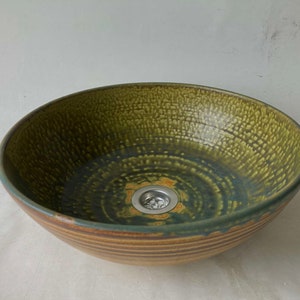 Può includere: Un lavandino in ceramica verde con un bordo marrone e uno scarico al centro. Il lavandino ha una superficie testurizzata e una forma leggermente arrotondata.