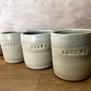 May include: Three ceramic mugs with personalised names, "Carresa", "Sylvi", and "Lorenz", in a light grey colour.
