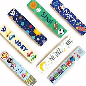 May include: Six personalized wooden mezuzahs with different designs. The designs include a space theme, a car theme, a superhero theme, a train theme, a sports theme, and a star theme. Each mezuzah has a Hebrew letter Shin on it and a name written in Hebrew and English.