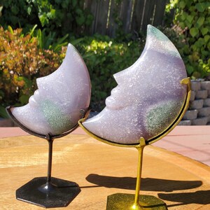 May include: Two crescent moon-shaped amethyst crystal sculptures on metal stands. The sculpture on the left has a black stand, and the sculpture on the right has a gold stand.