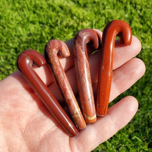 Puede incluir: Cuatro varitas de cristal de jaspe rojo con un mango curvo. Las varitas están pulidas y tienen un patrón natural de mármol rojo y blanco.