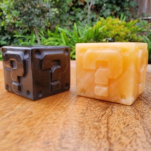 May include: Two question mark blocks, one black and one yellow, are on a wooden surface. The blocks are made of a solid material and have a 3D question mark design.