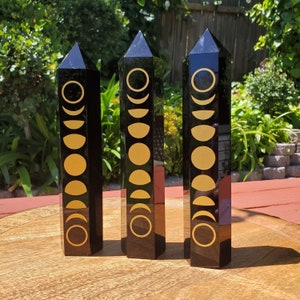 May include: Three black obsidian crystal towers with gold moon phases etched on each side. The towers are standing on a wooden surface.