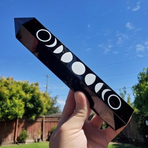 May include: Black obsidian crystal point with a white moon phase design etched on the surface. The crystal is approximately 6 inches long.