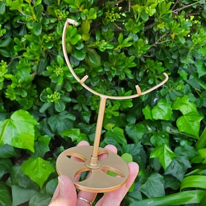 以下が含まれることがあります： 円形のベースと湾曲した上部を備えた、金色の金属製ディスプレイスタンド。スタンドはベースに十字型のデザインがあり、緑の葉を背景に手で持たれています。スタンドはアイテムを保持するように設計されています。