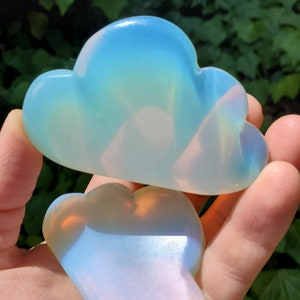 May include: Two iridescent opal cloud-shaped gua sha tools. The tools are made of smooth, polished opal and have a light blue and white color with a hint of pink.