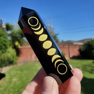 May include: Black obsidian crystal point with a gold moon phase design. The crystal is polished and has a smooth, shiny surface.