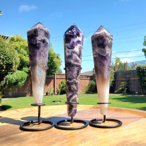 May include: Three amethyst crystal points, each mounted on a black metal stand. The crystals display a gradient of purple and white hues. The stands are circular at the base and have a ring to hold the crystal.