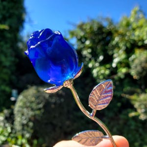 以下が含まれることがあります： メタリックゴールドの茎と葉を持つ鮮やかな青いバラの彫刻。バラは深く半透明の青色で、葉は詳細なテクスチャパターンを持っています。背景は、緑の葉と澄んだ青い空のぼやけた景色です。