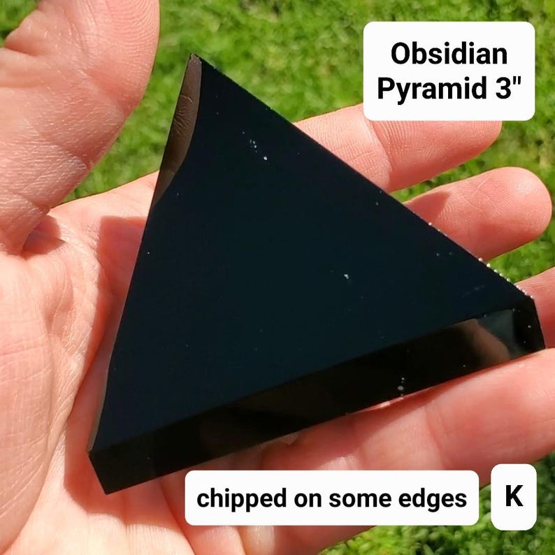 May include: A black obsidian pyramid, approximately 3 inches in height, with a triangular shape. The image shows the pyramid held in a hand against a green background. Text on the image reads "Obsidian Pyramid 3"" and "chipped on some edges."