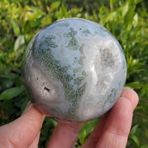 May include: A green and white agate sphere with a druzy finish. The sphere has a natural, organic look with a variety of textures and patterns.