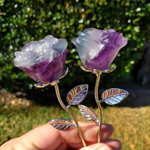 May include: Two carved fluorite roses featuring translucent purple and white color gradients. Each stone flower is attached to a polished silver-tone metal stem with two textured metal leaves, creating a pair of decorative floral sculptures.