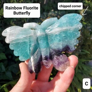 May include: A carved rainbow fluorite butterfly sculpture. The butterfly features a mix of colors, including shades of blue, purple, and white. The text "Rainbow Fluorite Butterfly" is displayed in a white box. The corner of the butterfly wing is chipped.