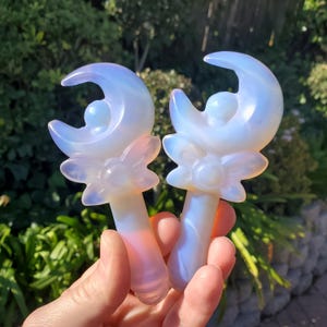 Può includere: Due bacchette di opale iridescente con un design a forma di luna crescente e fiore. Le bacchette sono bianche con un bagliore arcobaleno.