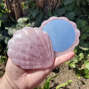May include: A light pink, translucent, shell-shaped compact mirror with a carved, ribbed exterior texture. The object consists of two hinged pieces, with one side featuring a circular reflective glass surface and the other a decorative stone shell.