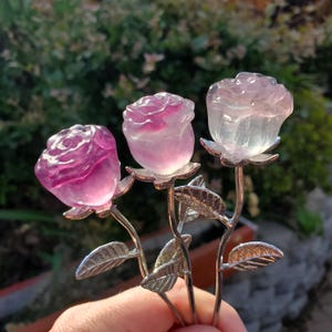 May include: Three crystal rose sculptures in shades of pink and clear. Each rose is mounted on a silver-coloured stem with detailed leaves. The roses are held in front of a blurred green background.
