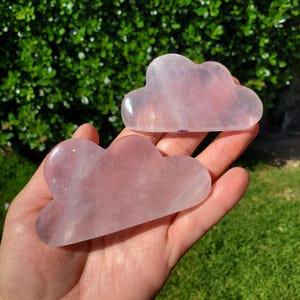 May include: Two pink rose quartz cloud-shaped massage tools. The tools are held in a hand, with a green leafy background. The rose quartz has a translucent quality and a soft pink hue. These tools are used for facial massage and skincare.