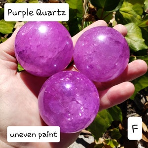 May include: Three polished purple quartz spheres. The spheres are held in a hand, with the text "Purple Quartz" and "uneven paint" visible. The spheres have a translucent appearance.