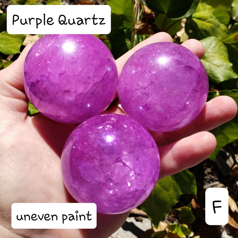 May include: Three polished purple quartz spheres. The spheres are held in a hand, with the text "Purple Quartz" and "uneven paint" visible. The spheres have a translucent appearance.