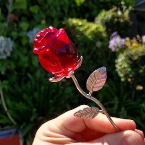 Puede incluir: Una escultura de rosa roja con tallo y hojas plateadas. La rosa es de un rojo profundo y translúcido, y las hojas tienen un diseño texturizado detallado. La escultura se sostiene en una mano sobre un fondo verde borroso.
