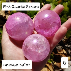 May include: Three pink quartz spheres are held in a hand. The spheres have a translucent appearance with internal crystalline structures. The text "Pink Quartz Sphere" and "uneven paint" are visible. The spheres are a vibrant pink color.